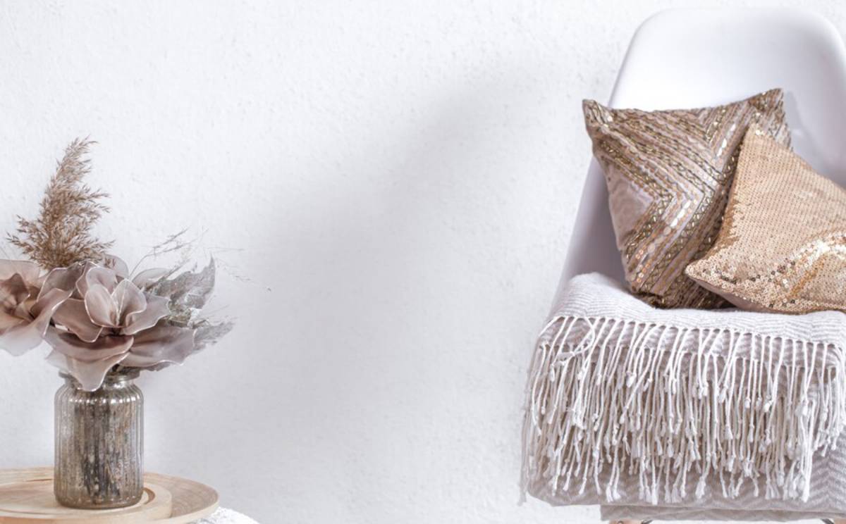 Cozy corner featuring a white chair with decorative pillows, a fringed throw, and a glass vase of muted faux flowers and dried grass.