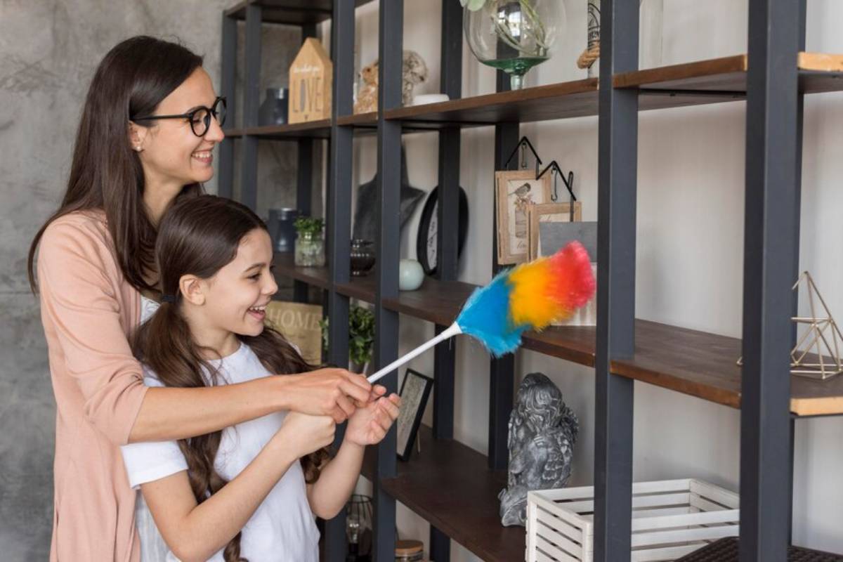 A woman assists a girl with dusting a shelf, surrounded by decorative objects and plants in a modern living space.