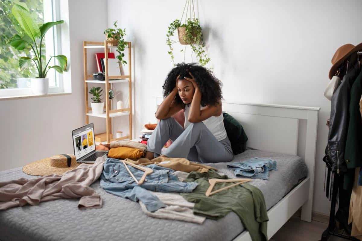 A person sits on a bed surrounded by a messy pile of clothes, with a laptop displaying fashion items nearby.