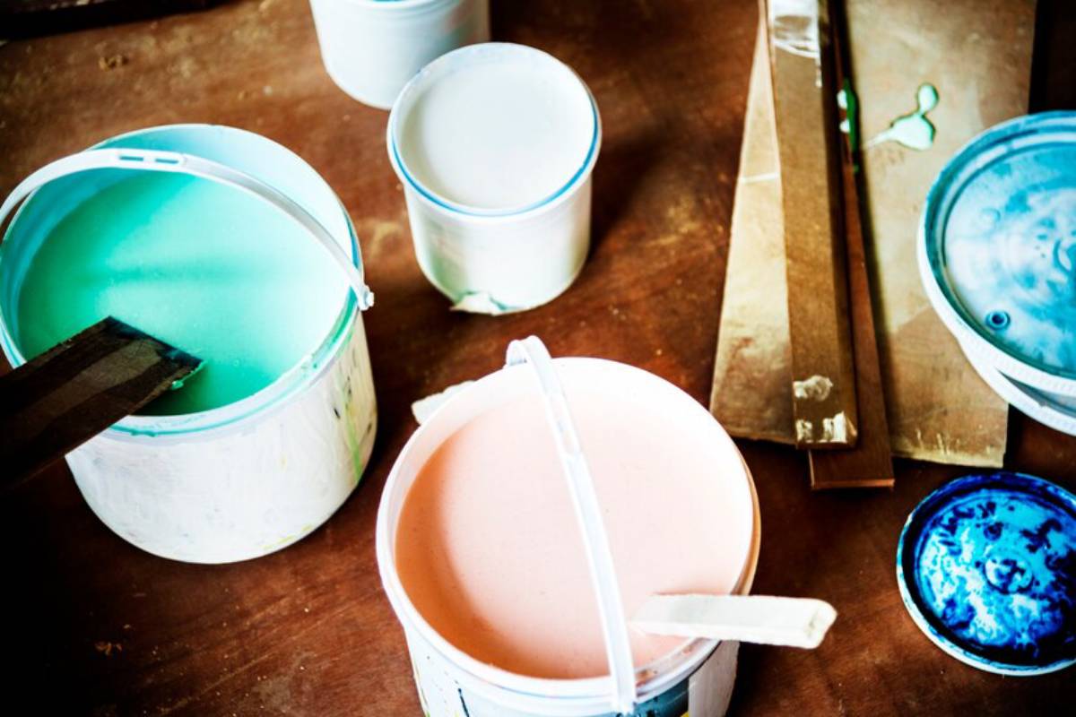 Various paint cans with green, pink, and blue paint sit on a wooden surface, alongside mixing sticks and palette boards.